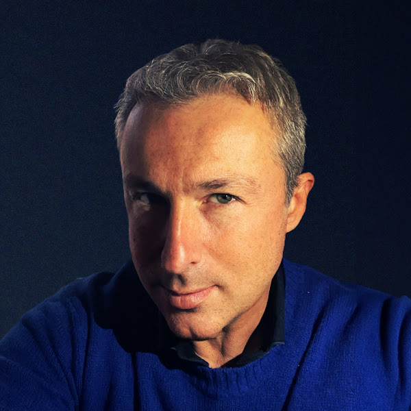 Paolo Chiesa con capelli grigi corti e maglione blu, sguardo diretto, sfondo scuro, illuminazione laterale.